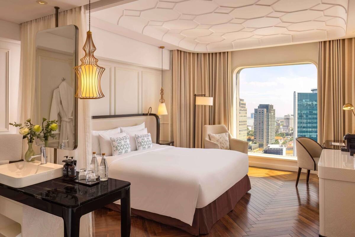 A modern hotel room with a large bed, a desk with a chair, a vanity with a mirror, and a large window offering a city view.