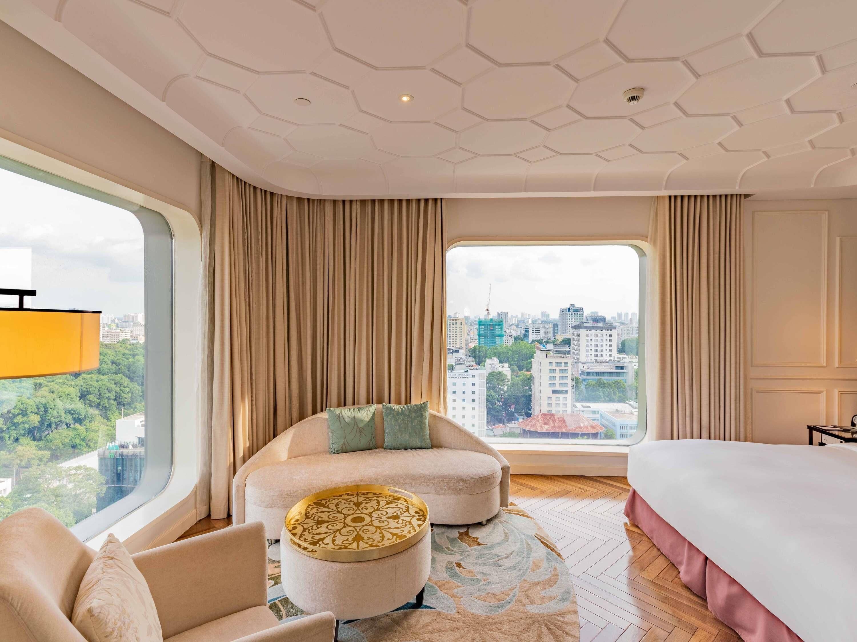 A hotel room with a large window, a bed, a sofa, a round ottoman, and a view of a cityscape.