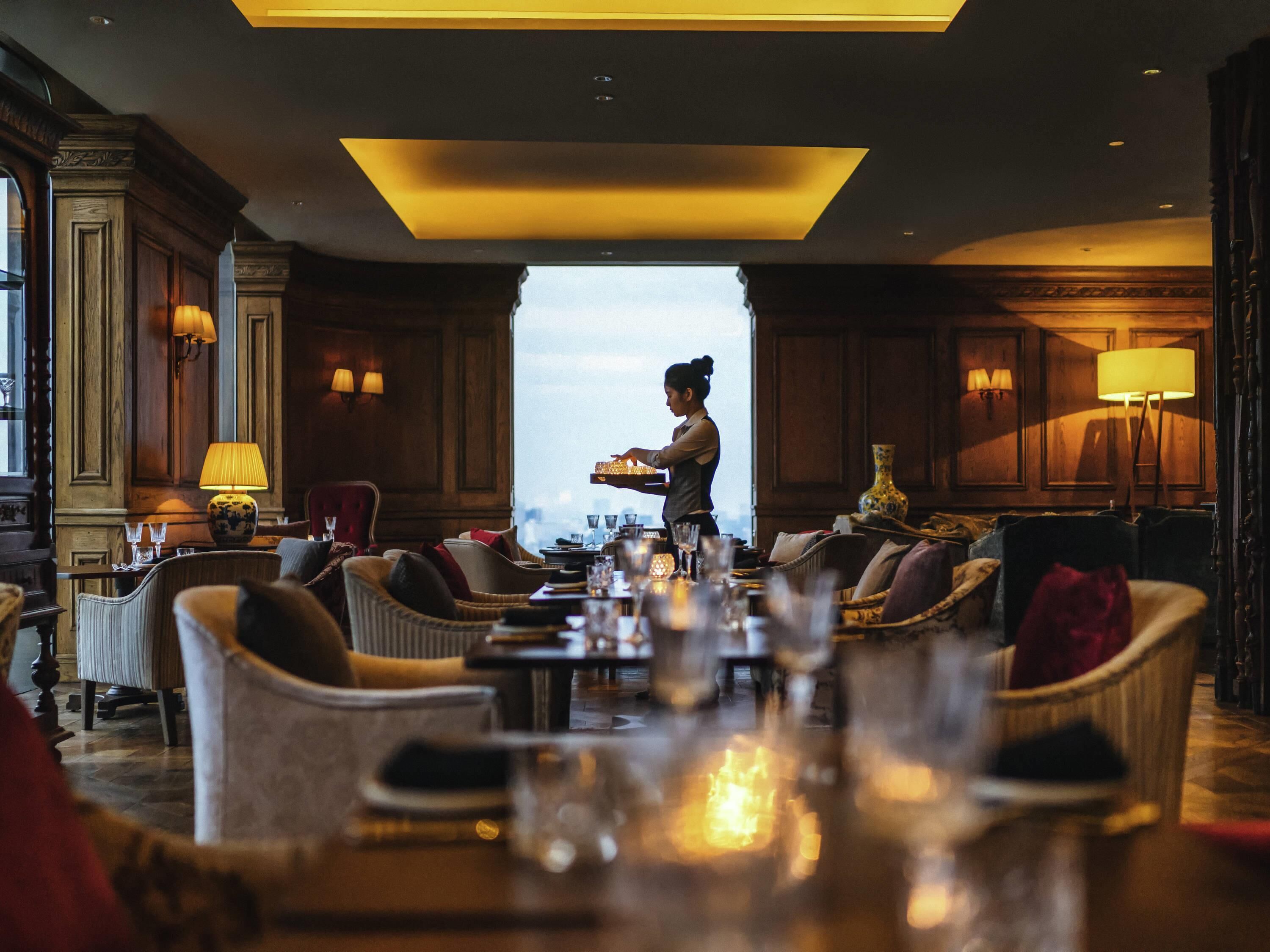 A spacious hotel lobby with a central seating area, a waiter attending to guests, and a view through a large window.