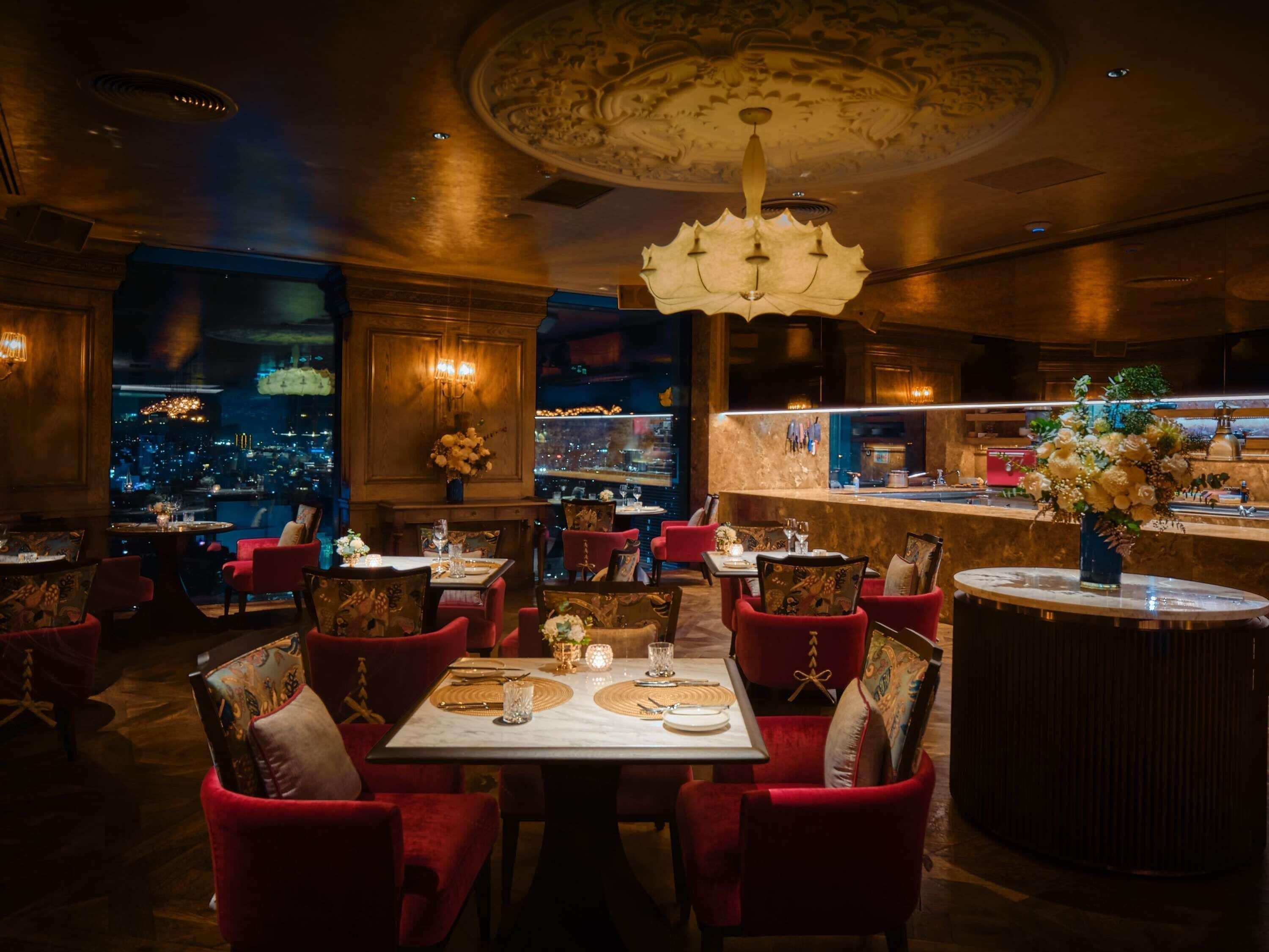 A restaurant with a city view, red upholstered chairs, a marble table, and a chandelier.