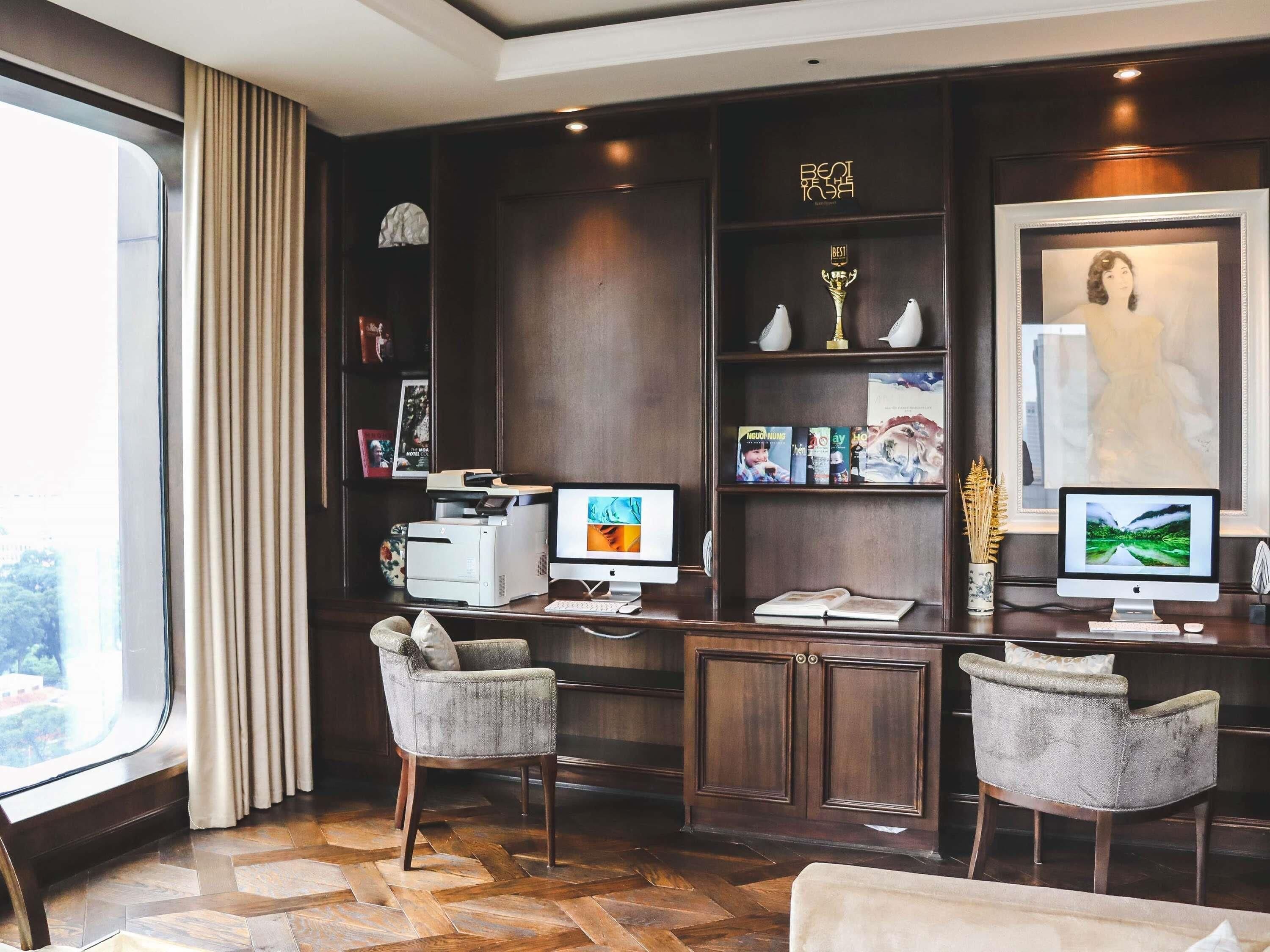 A hotel room with a desk, two chairs, a printer, a computer, a bookshelf with books and trophies, and a large window with curtains.