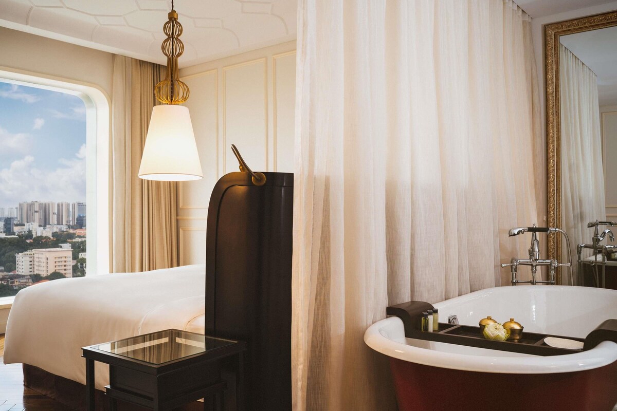 Une chambre d’hôtel avec un lit, une table en verre, une grande fenêtre, une salle de bain avec baignoire et une vue sur la ville.