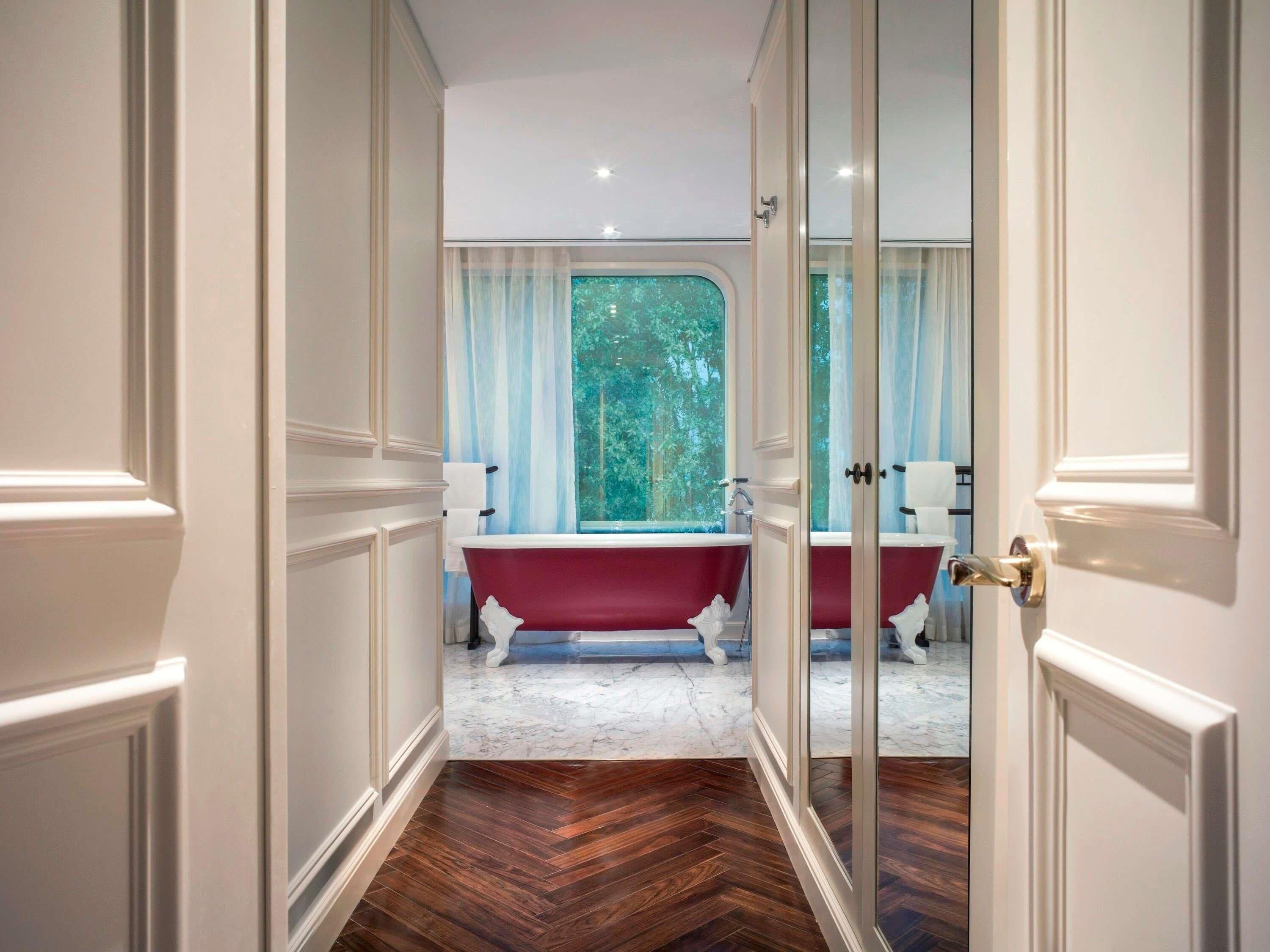 Un baño con una bañera roja, accesorios blancos y vistas a árboles por una ventana.