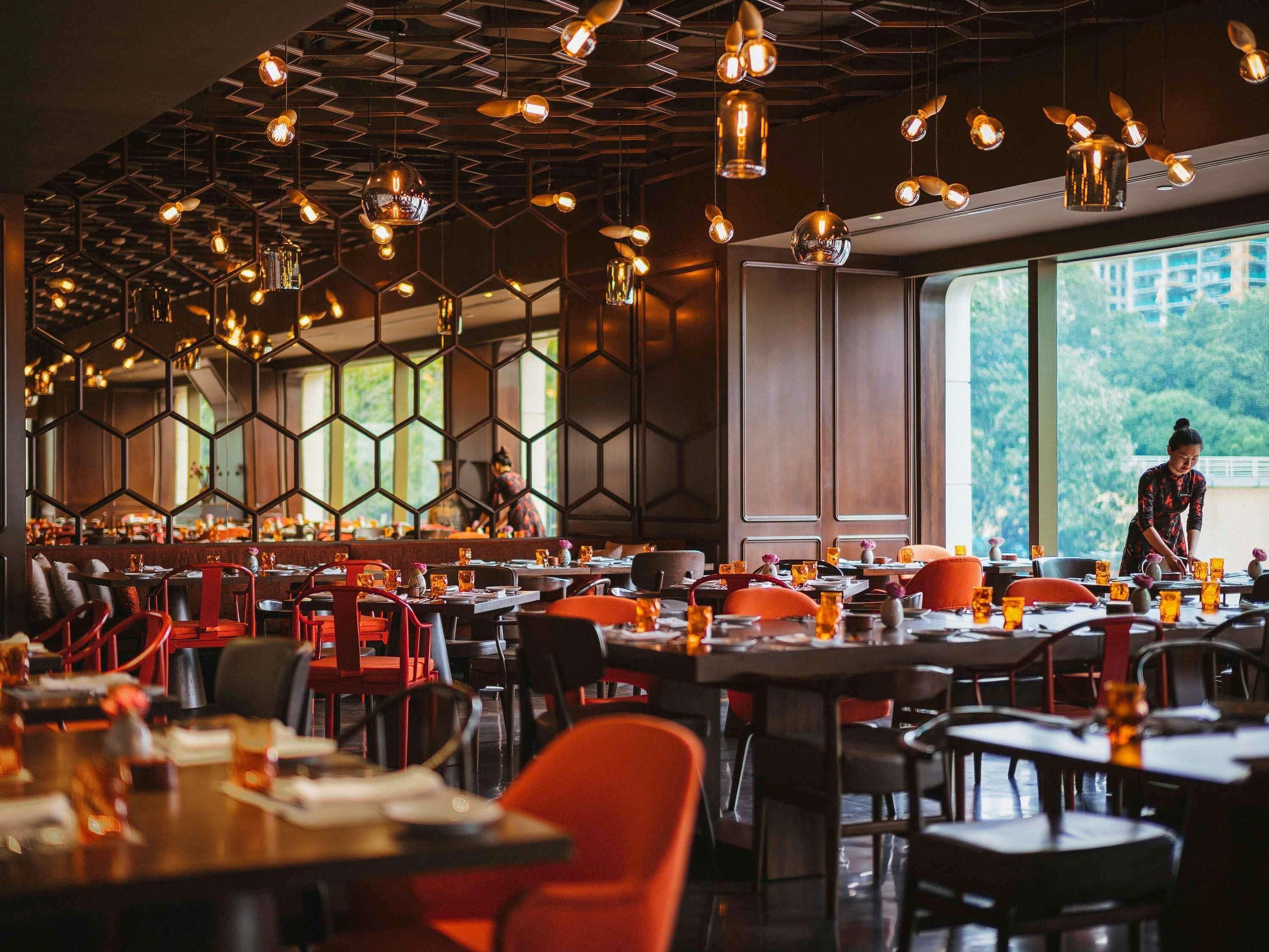A restaurant interior with hexagonal ceiling panels, tables with orange chairs, and a view of a person working in the background.