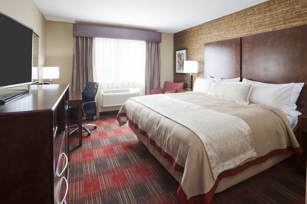 Ein Hotelzimmer mit einem großen Bett, einem Schreibtisch mit Fernseher, einem Sessel und einem Fenster mit Vorhängen.