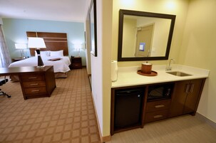 Habitación de hotel con cama, mesitas de noche, escritorio, espejo, lavabo y lavavajillas integrado.