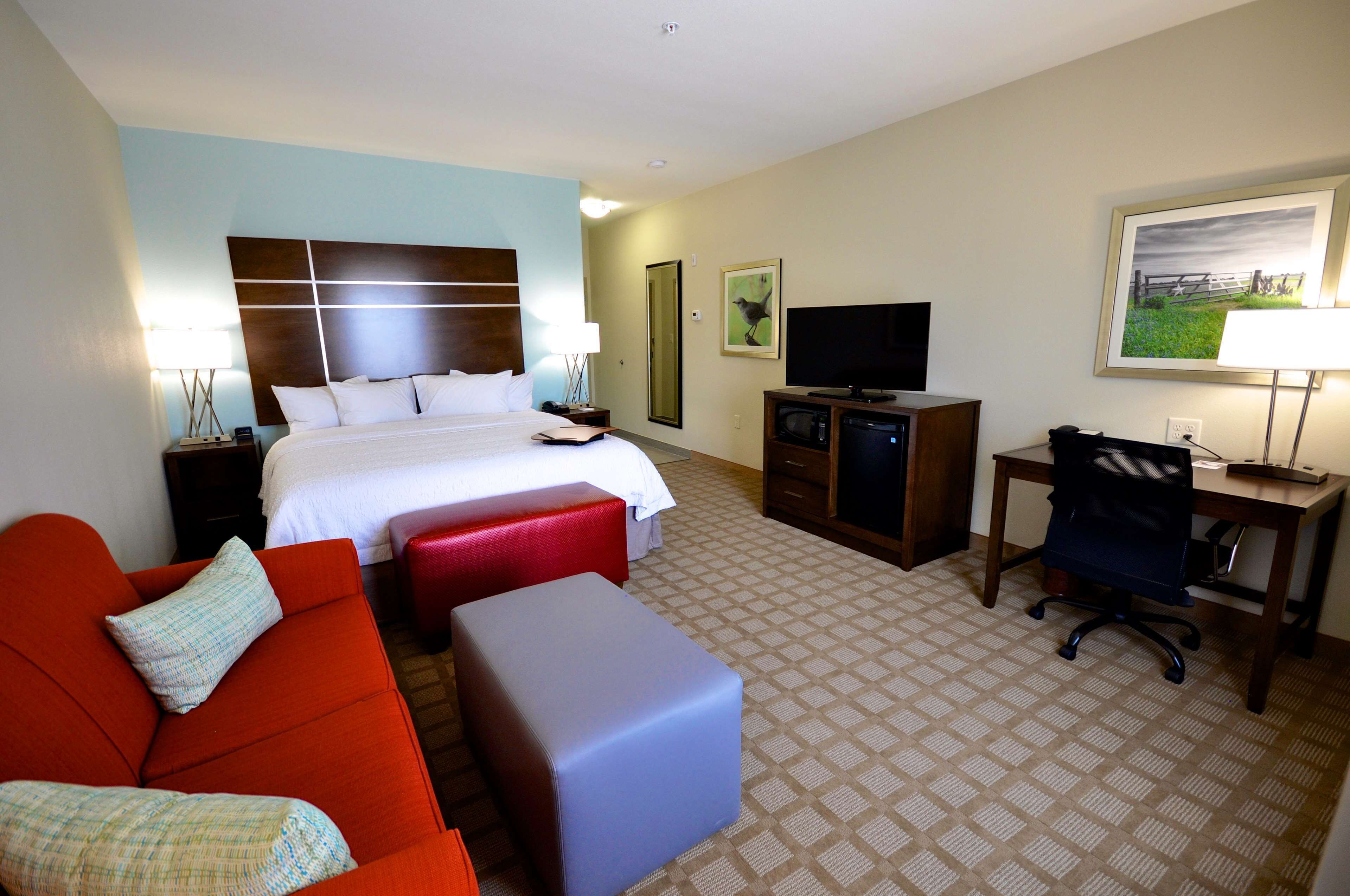 A hotel room with a bed, a red sofa, a blue ottoman, a desk with a chair, a television, and artwork on the walls.
