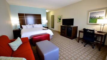 A hotel room with a bed, a red sofa, a blue ottoman, a desk with a chair, a television, and artwork on the walls.