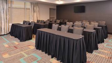 Un salón de conferencias con mesas y sillas, un ventanal con cortinas y un televisor de pantalla plana en la pared.