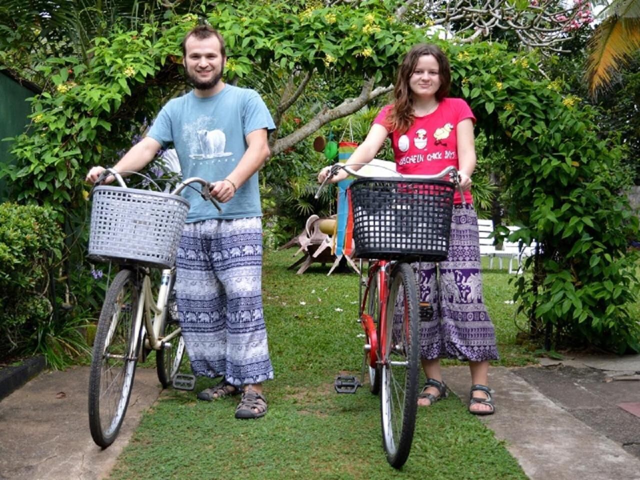 Duas pessoas de bicicleta, com cestas, em uma área gramada, com vegetação ao fundo.