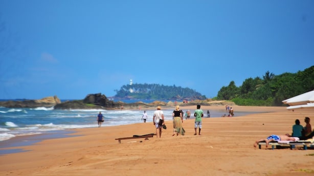 A beach with people walking and sitting, a rocky shoreline, and a lighthouse in the distance.