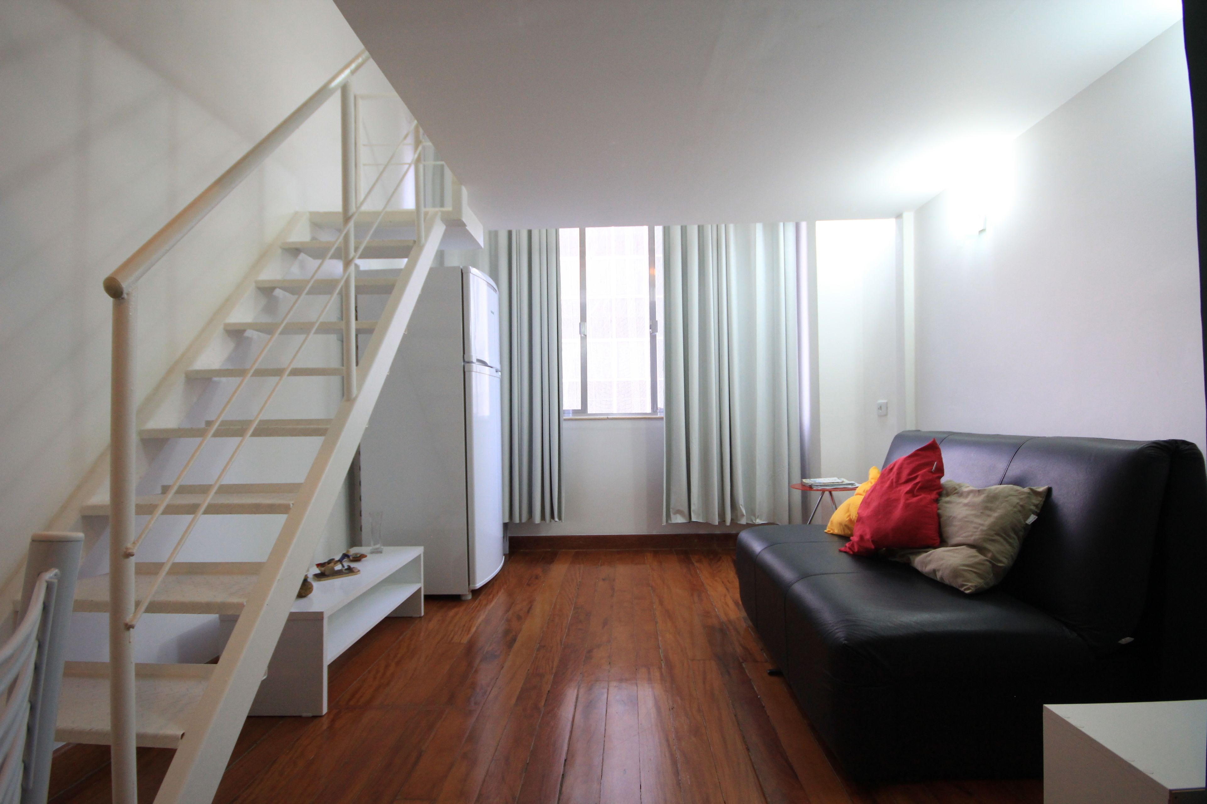 A living room with a staircase, a black sofa with cushions, a white refrigerator, and a window with curtains.
