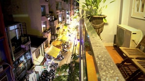 Vista nocturna de una calle estrecha flanqueada por edificios de varios pisos, con un balcón que tiene una planta en maceta y una silla.