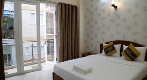Habitación de hotel con cama, mesita de noche, lámpara de pared, ventana con cortinas y vista al balcón exterior.