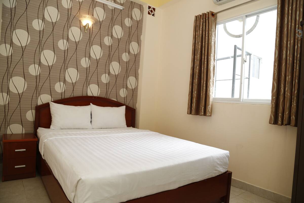 A neatly made bed with white linens in a room with a patterned wallpaper, a nightstand, and a window with curtains.