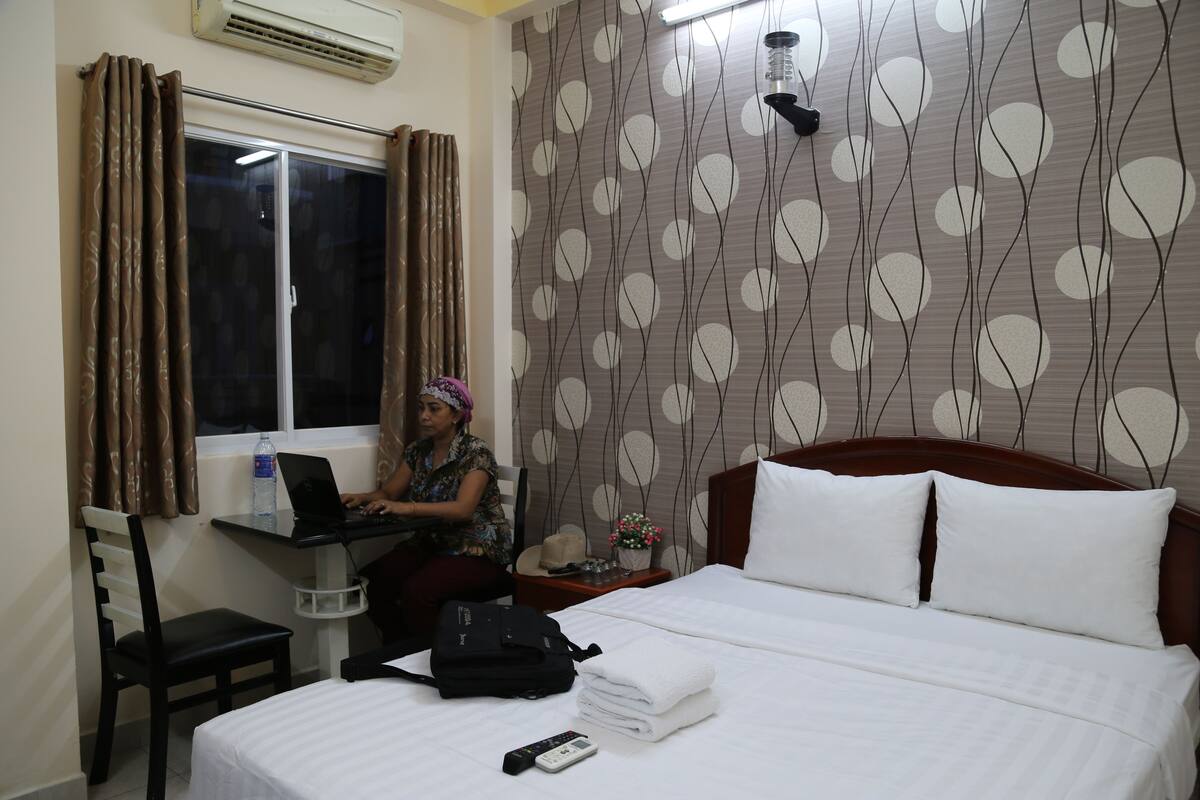 A hotel room with a bed, a desk with a laptop, a chair, a suitcase, and a window with curtains.