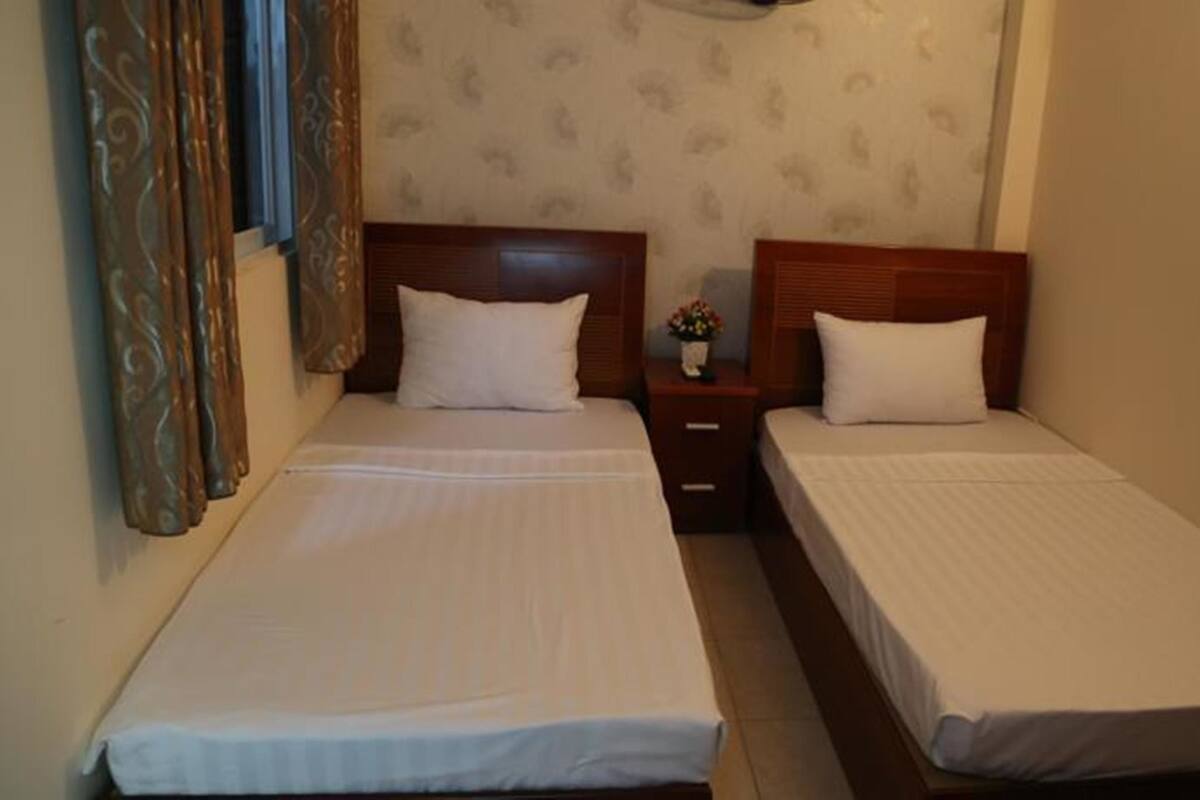 A room with two single beds, each with a wooden headboard and white bedding. There is a small bedside table with a vase of flowers between the beds.