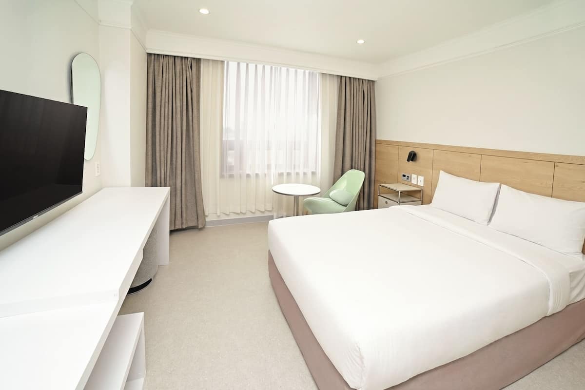 Una habitación de hotel con cama, televisión, escritorio, silla, mesita y cortinas.