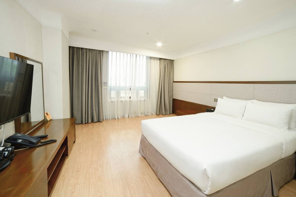 Habitación de hotel con cama, televisor, escritorio con teléfono y ventana con cortinas.