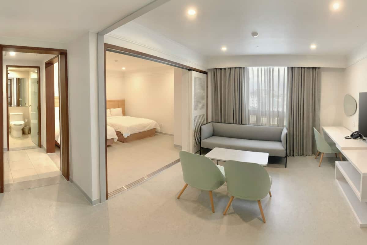 Habitación de hotel moderna con cama, sofá, escritorio con silla, televisor y baño visible a través de una puerta abierta.