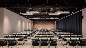 A conference room with rows of chairs and tables, a stage with a curtain, and a ceiling with exposed lighting and ductwork. - Staz Hotel Heritage (Jeju City)