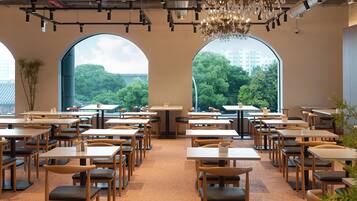 A restaurant interior with multiple tables and chairs, large arched windows offering a view of trees and buildings, and a chandelier hanging from the ceiling.