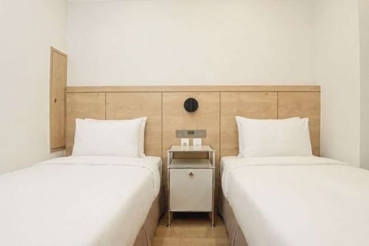 A hotel room with two beds, a wooden headboard, a nightstand with a clock, and a small table.
