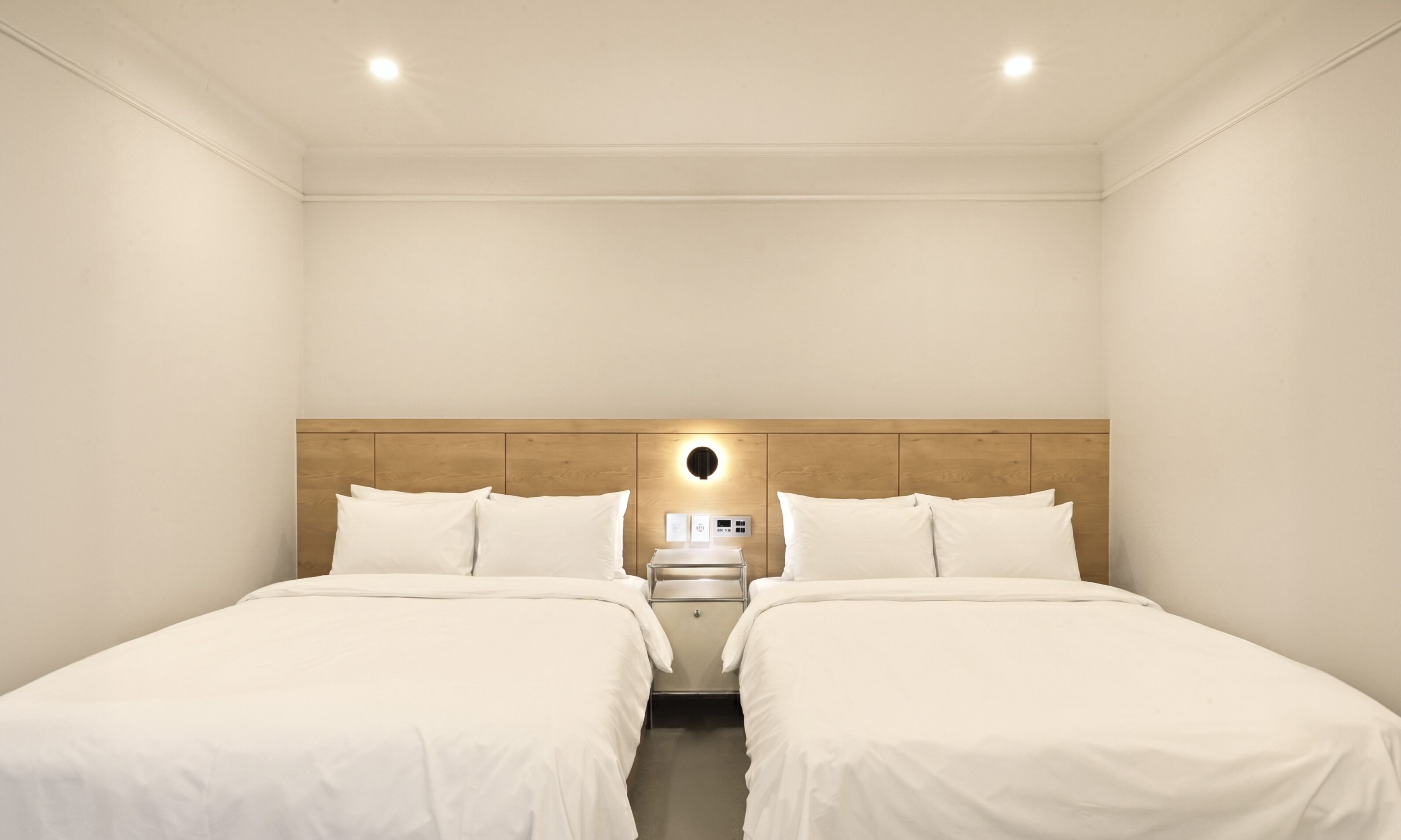 Habitación de hotel con dos camas, un cabecero de madera, una mesita de noche con lámpara y un panel de control.