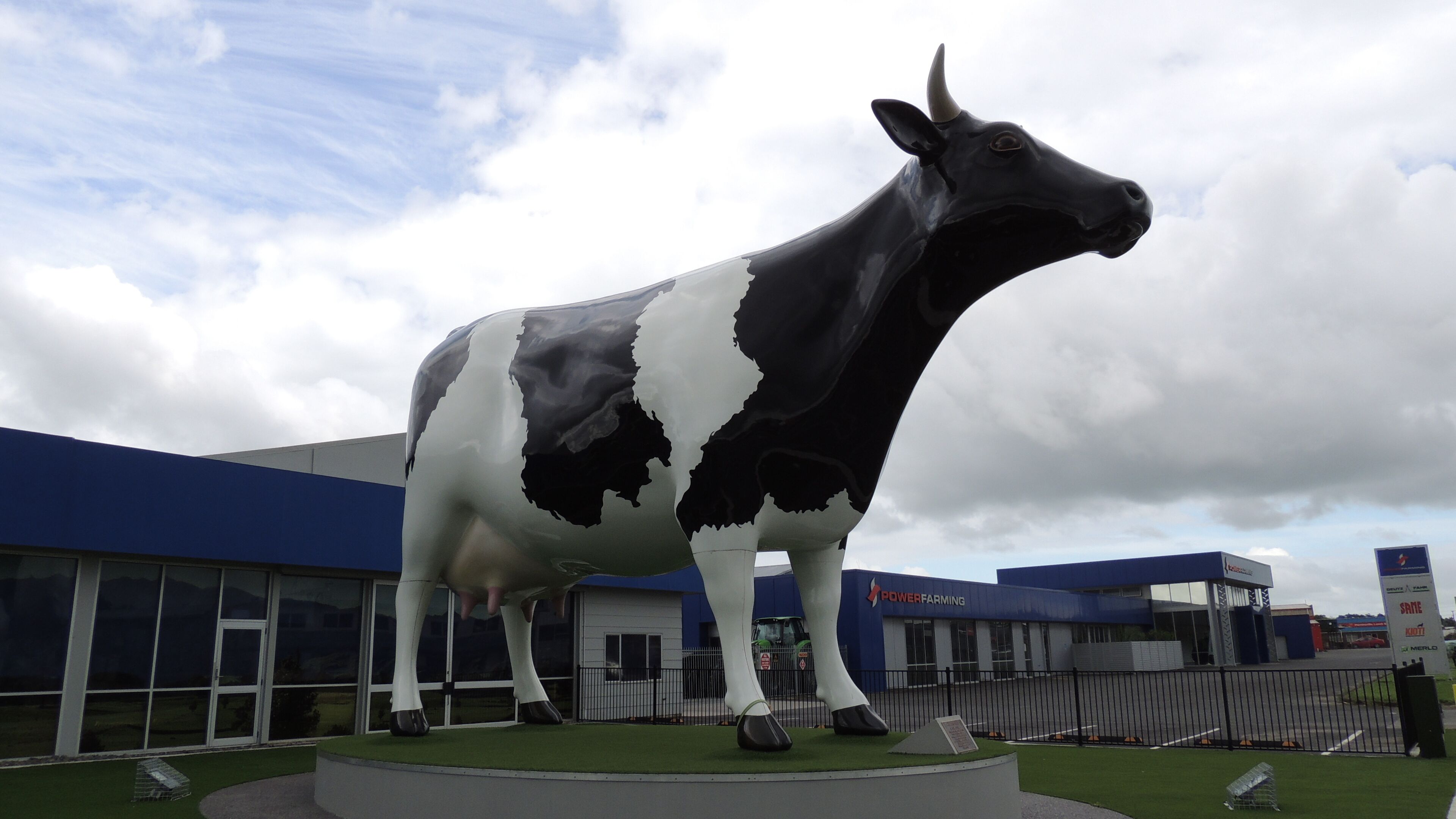 A large black and white cow statue in front of a building with a blue roof and a sign that reads "POWER FARMING".