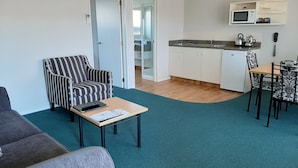 A compact living space with a striped armchair, a small table with a remote control, a kitchenette with a microwave, and a dining area with patterned chairs. - Central Motor Lodge (Morrinsville)