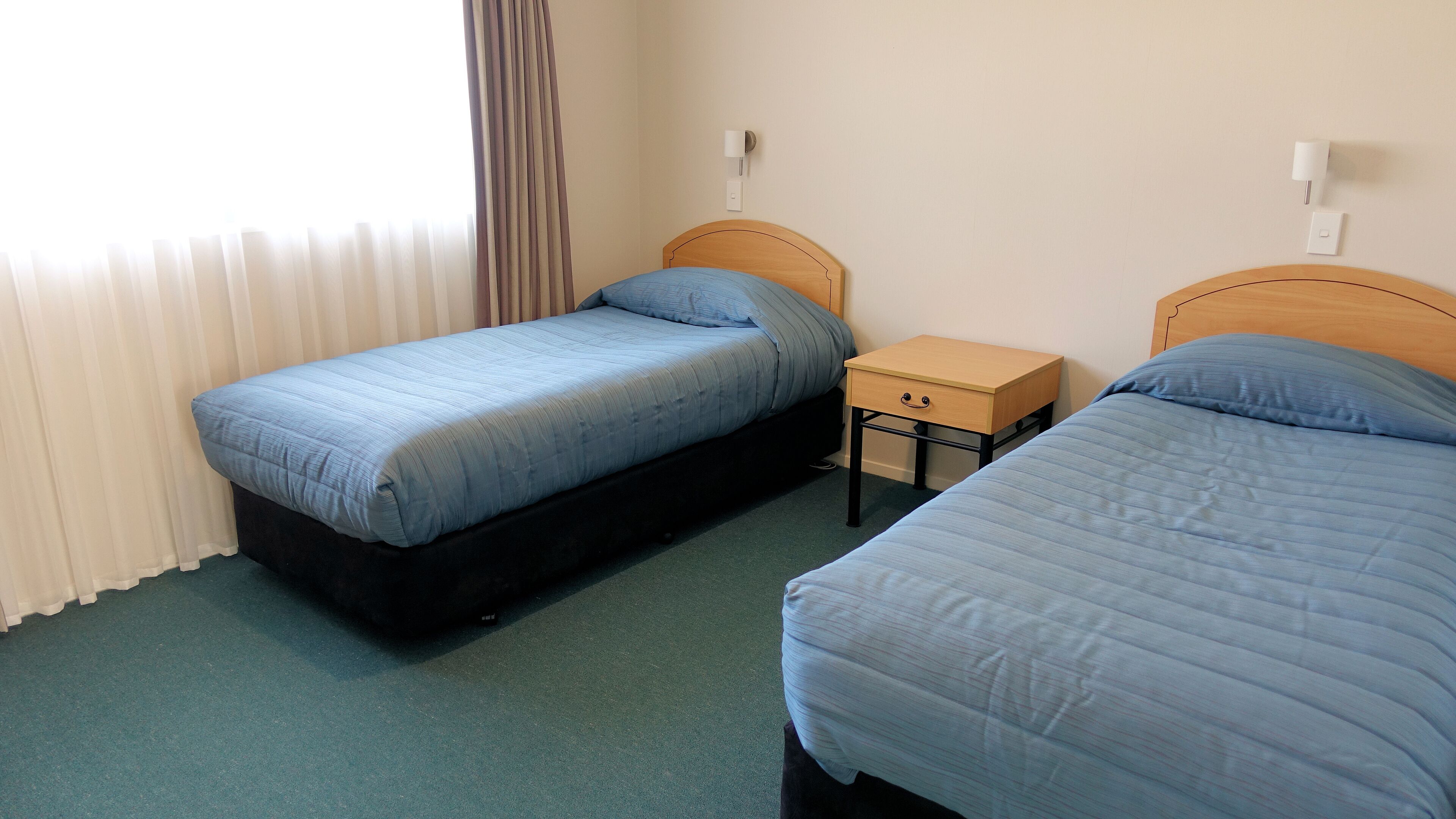 A hotel room with two single beds, a small wooden nightstand, and a window with curtains.