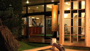 Ein traditionelles japanisches Onsen mit einer Holzwanne, einer Holzsäule mit Kalligraphie und Blick auf ein modernes Interieur mit Schiebetüren.