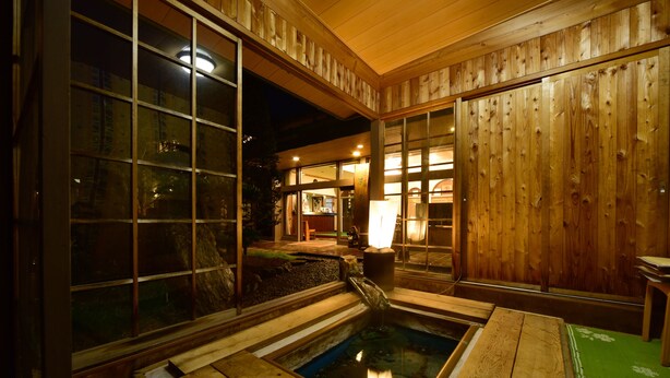 Ein traditionelles Holzbadhaus mit Holzinnenraum, einer kleinen Wasserspende und Blick ins Freie.