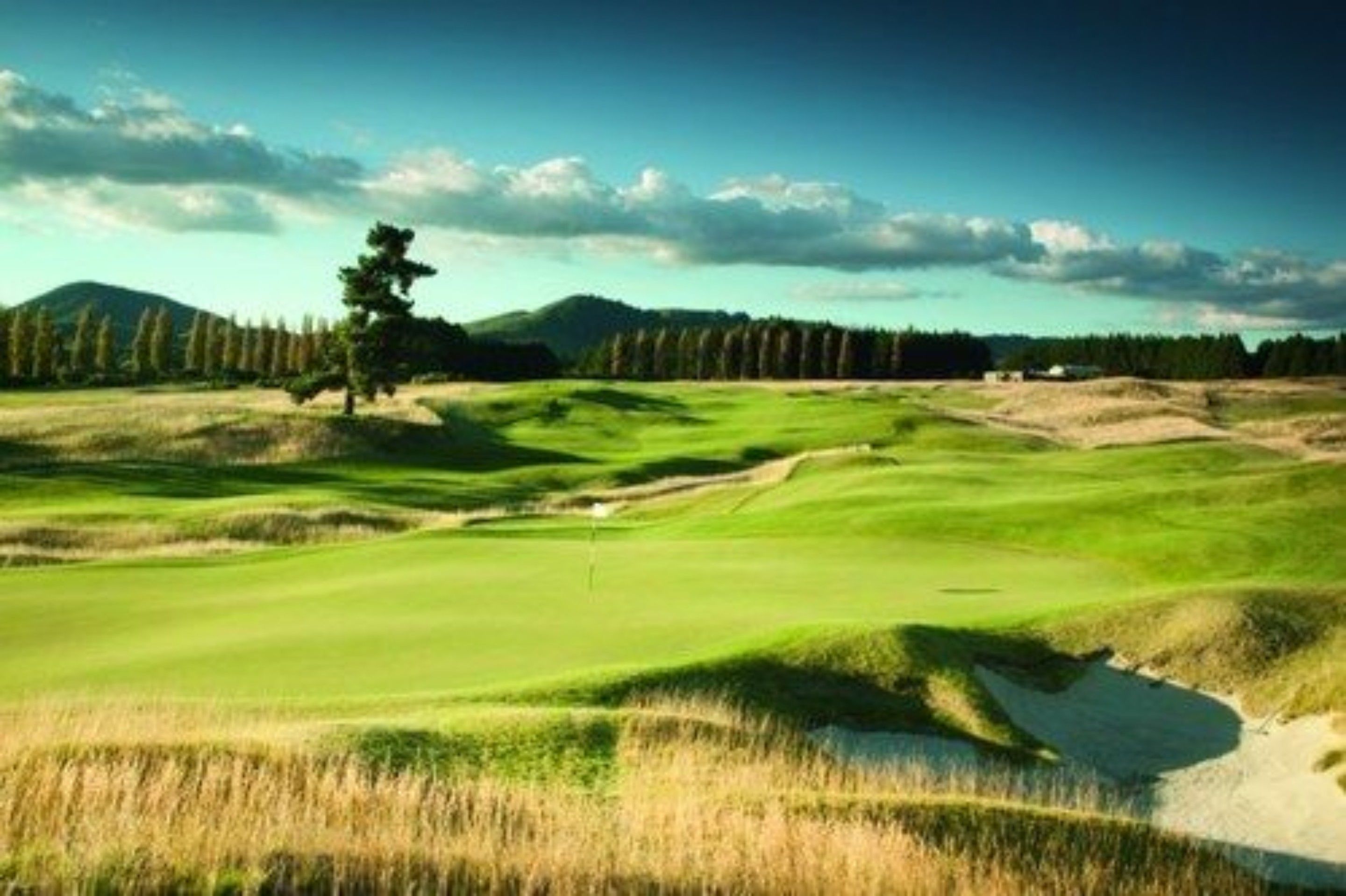 A golf course with a clear path, a flagstick, and a backdrop of trees and hills.