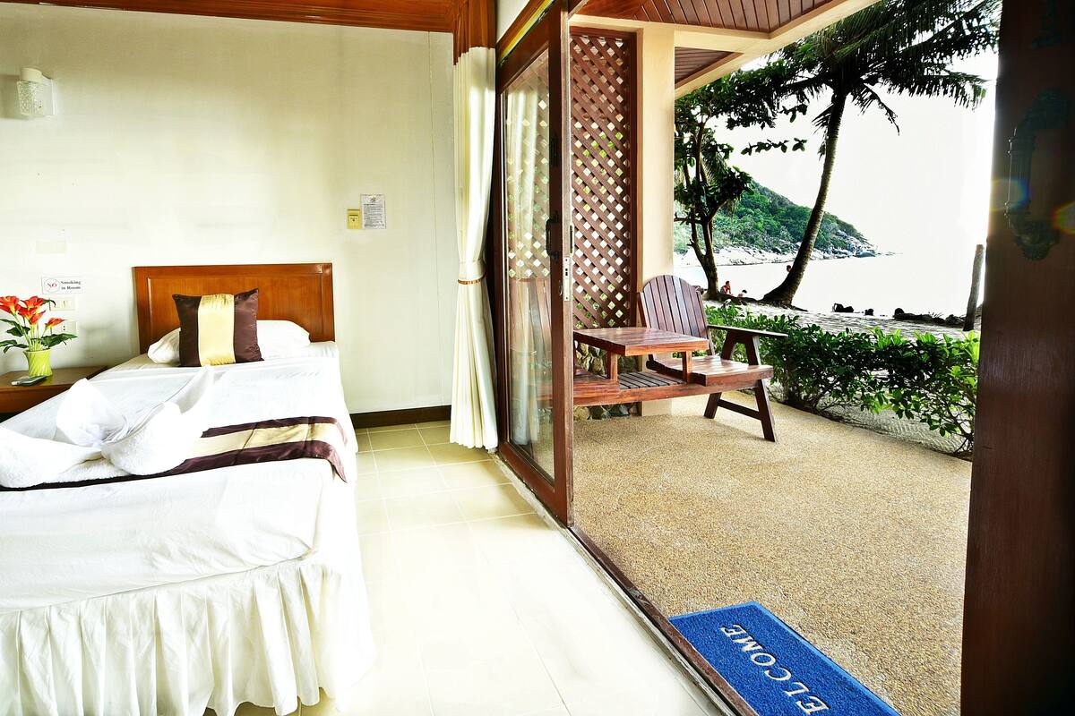 A hotel room with a bed, a small table with a vase of flowers, and a view of a beach and palm trees.