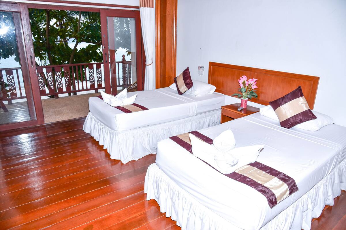 A hotel room with two beds, wooden flooring, a balcony with a view of trees, and a wooden headboard.