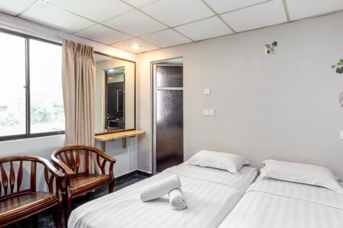 Habitación de hotel con dos camas, una silla de madera, una mesita, un espejo y una ventana con cortinas.