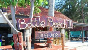 Un letrero de madera rústico con el texto "Bottle Beach II Jing Jing" en una tipografía rústica, sujeto a una estructura de ladrillo con barandilla metálica.
