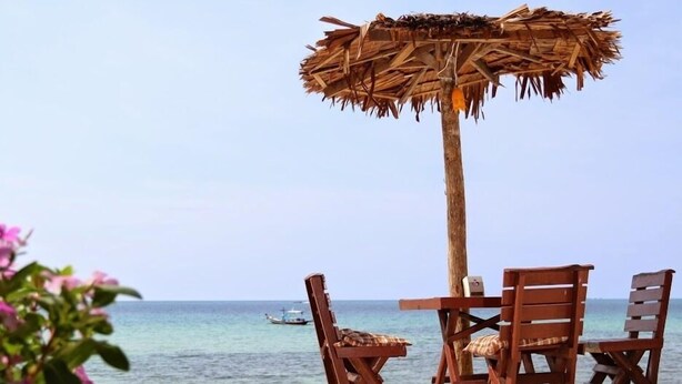 Un paisaje de playa con una mesa y sillas de madera bajo un toldo de paja, con vista al mar.