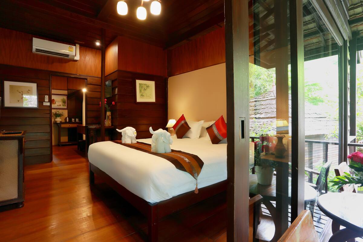 A hotel room with a wooden floor, a bed with white bedding, a desk with a lamp, and a view of the outdoors through a glass door.