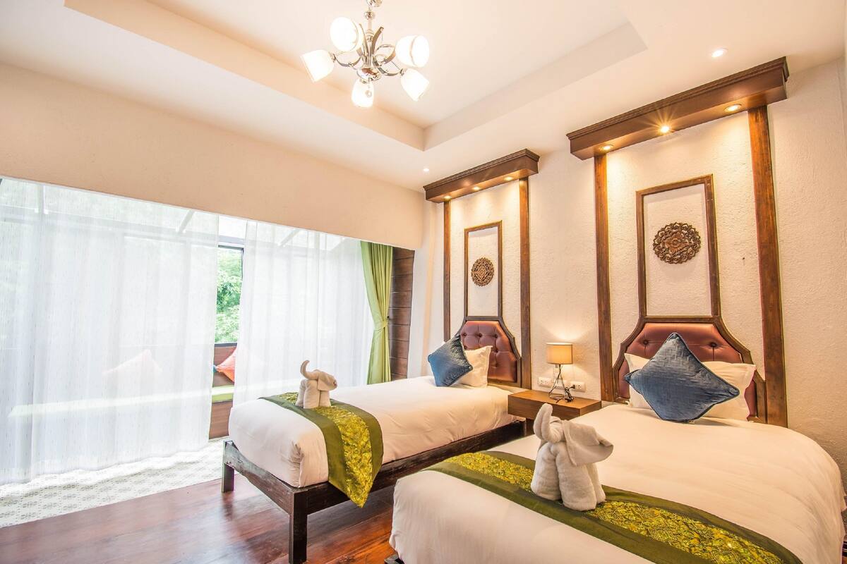 A hotel room with two beds, a chandelier, and a view of greenery outside.