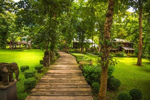 A wooden pathway leading through a lush garden with well-maintained grass, trees, and a small pond. - E-Outfitting Doikham Resort (Chiang Mai)