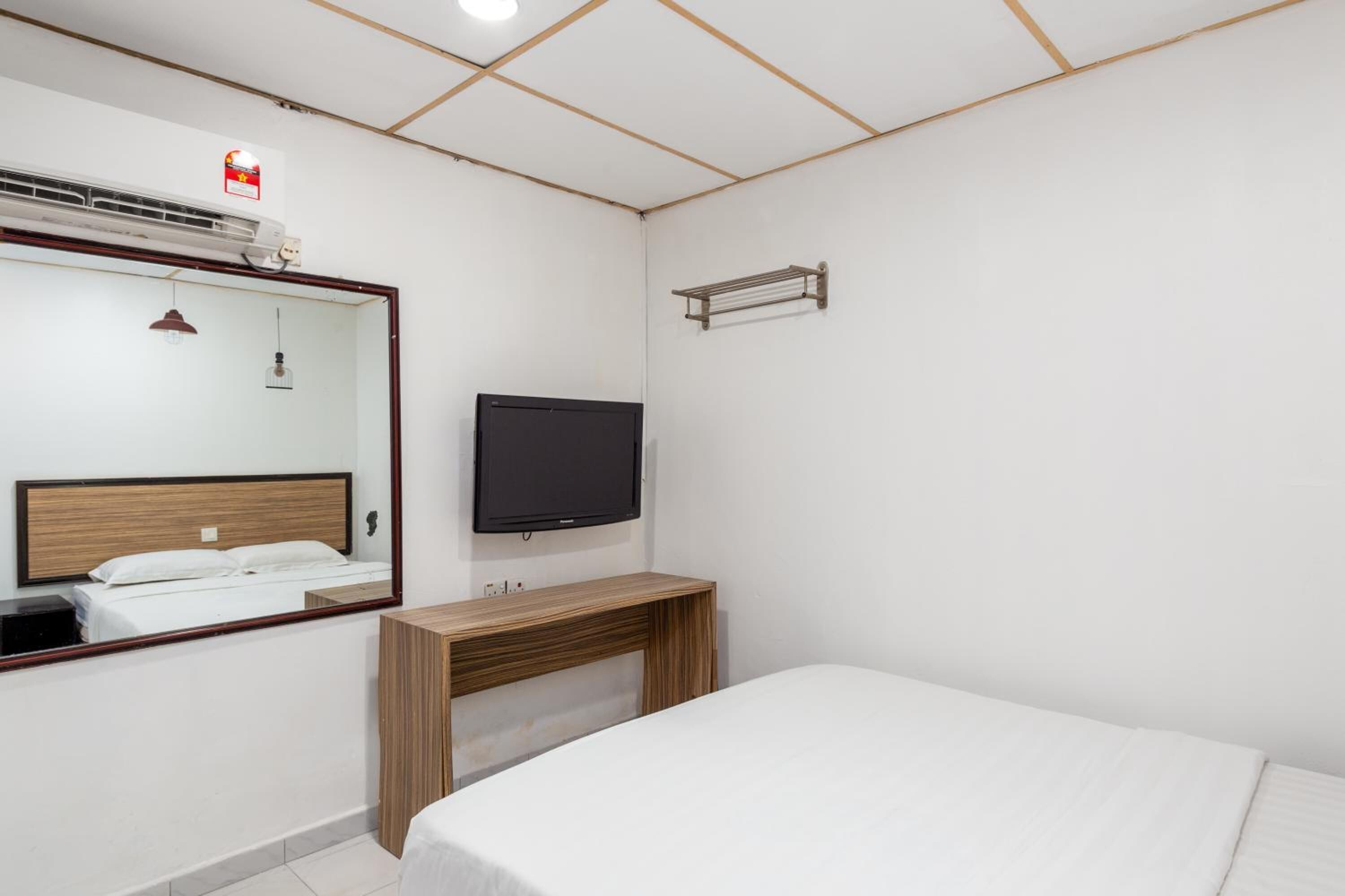 A hotel room with a bed, a TV, a wooden desk, a mirror, and a wall-mounted air conditioning unit.