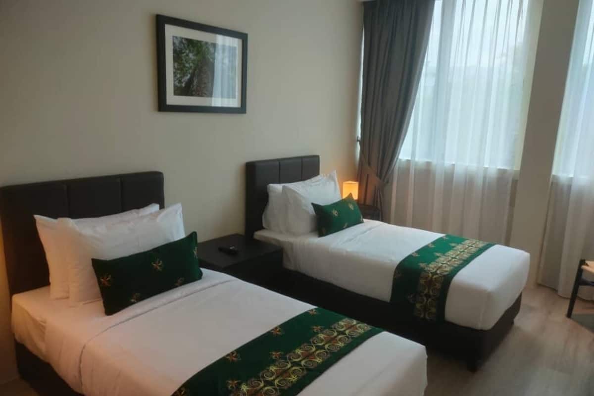 Habitación de hotel con dos camas, cada una con ropa blanca y almohadones verdes decorativos. Hay un cuadro en la pared y una ventana con cortinas.
