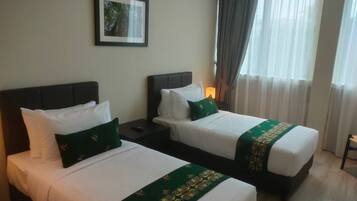 A hotel room with two beds, each with white linens and green decorative pillows. There is a framed picture on the wall and a window with curtains.