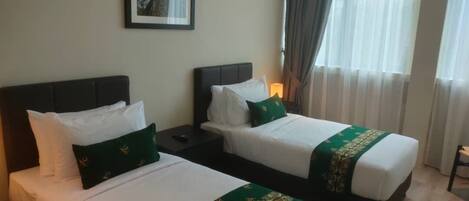 Habitación de hotel con dos camas, cada una con ropa blanca y almohadones verdes decorativos. Hay un cuadro en la pared y una ventana con cortinas.