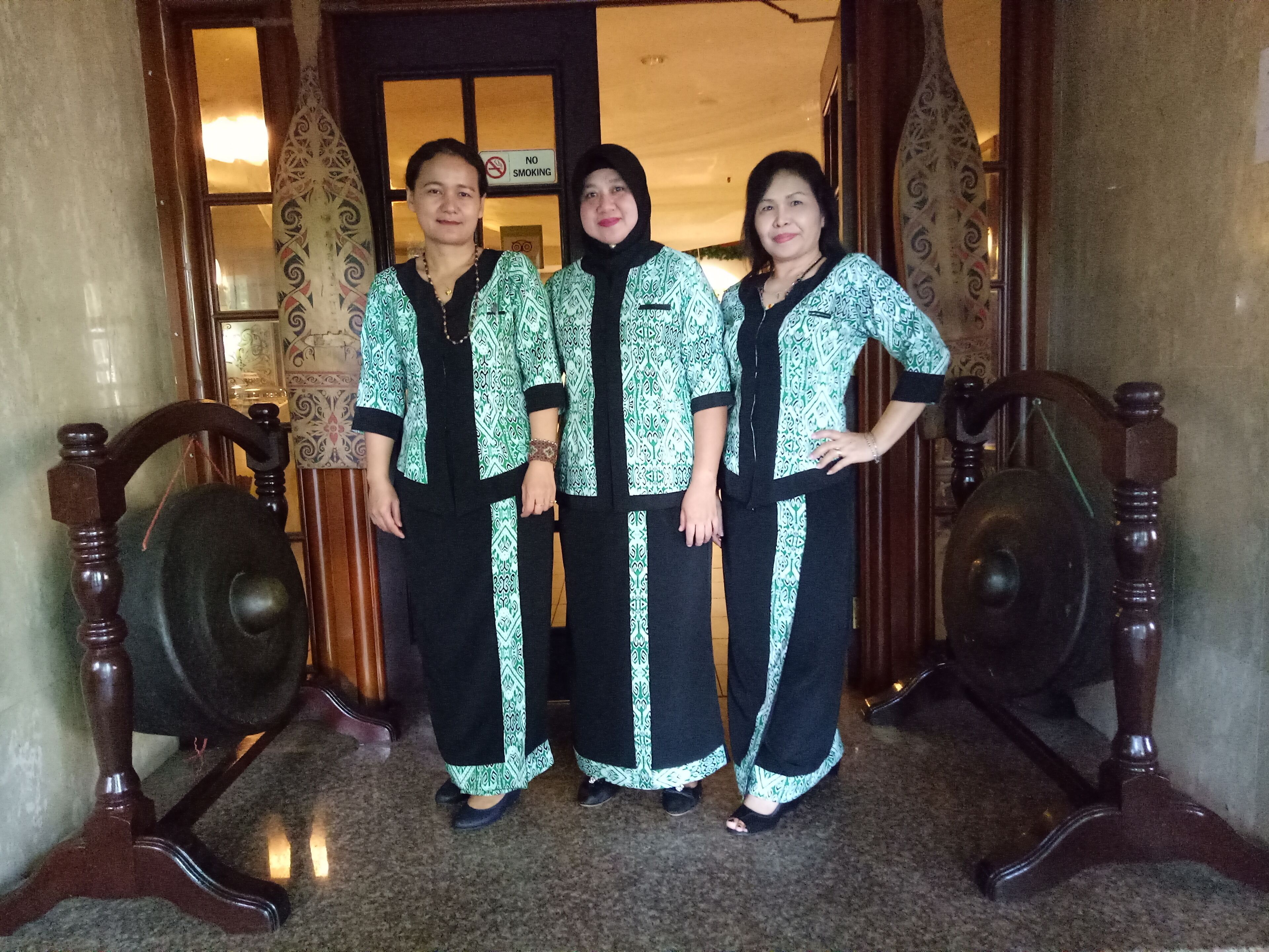 Three people standing in a doorway with patterned clothing, wooden furniture, and a 'No Smoking' sign.