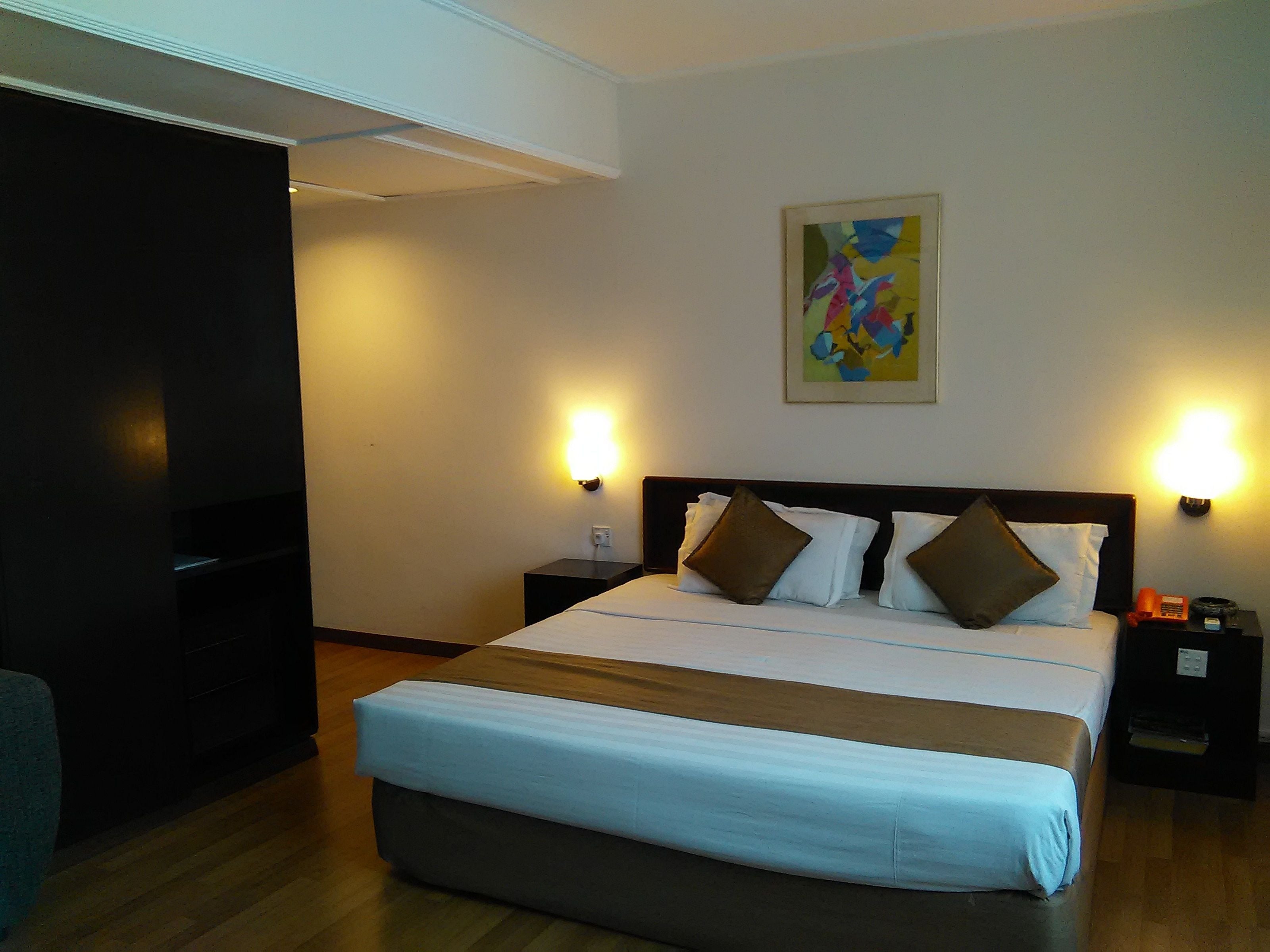 A hotel room with a large bed, bedside tables, a wall-mounted painting, and a television.