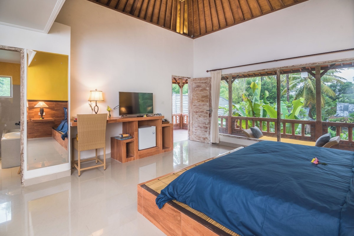 A bedroom with a large bed, a desk with a TV, and a view of lush greenery.