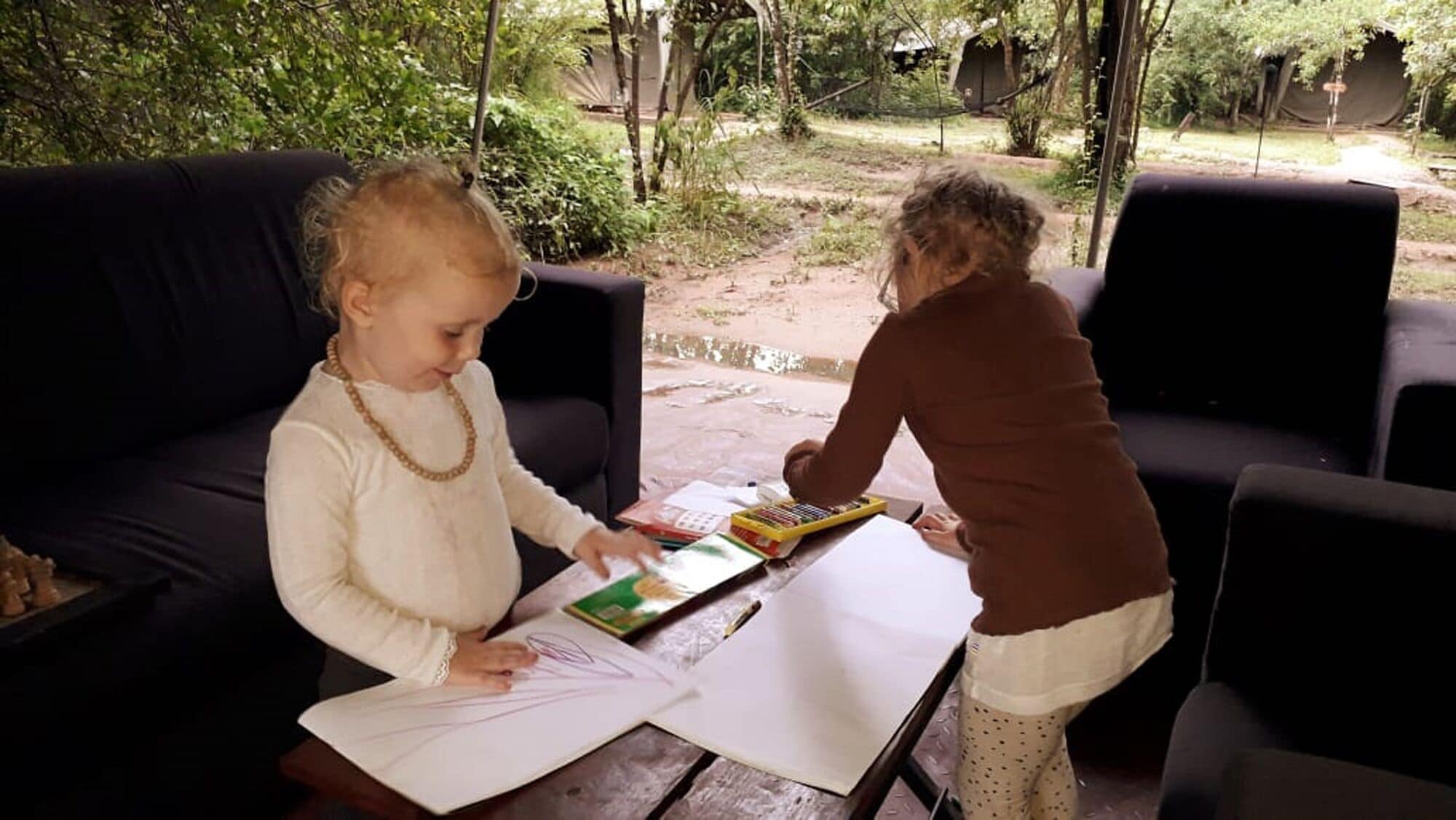Dos niños sentados a una mesa con libros y papeles, al aire libre con un jardín y una terraza de madera al fondo.